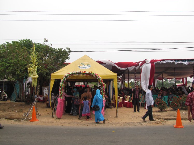 The welcoming archway as Despri and Nisa's wedding. Family members are waiting to greet guests under the yellow pop-up tent. The couple and their parents are sitting on a stage immediately to the left under the big tent, and rows and rows of chairs are set up facing the tent so that guests can people-watch while consuming massive amounts of food from the heavily laden buffet tables that line the perimeter of the big tent.